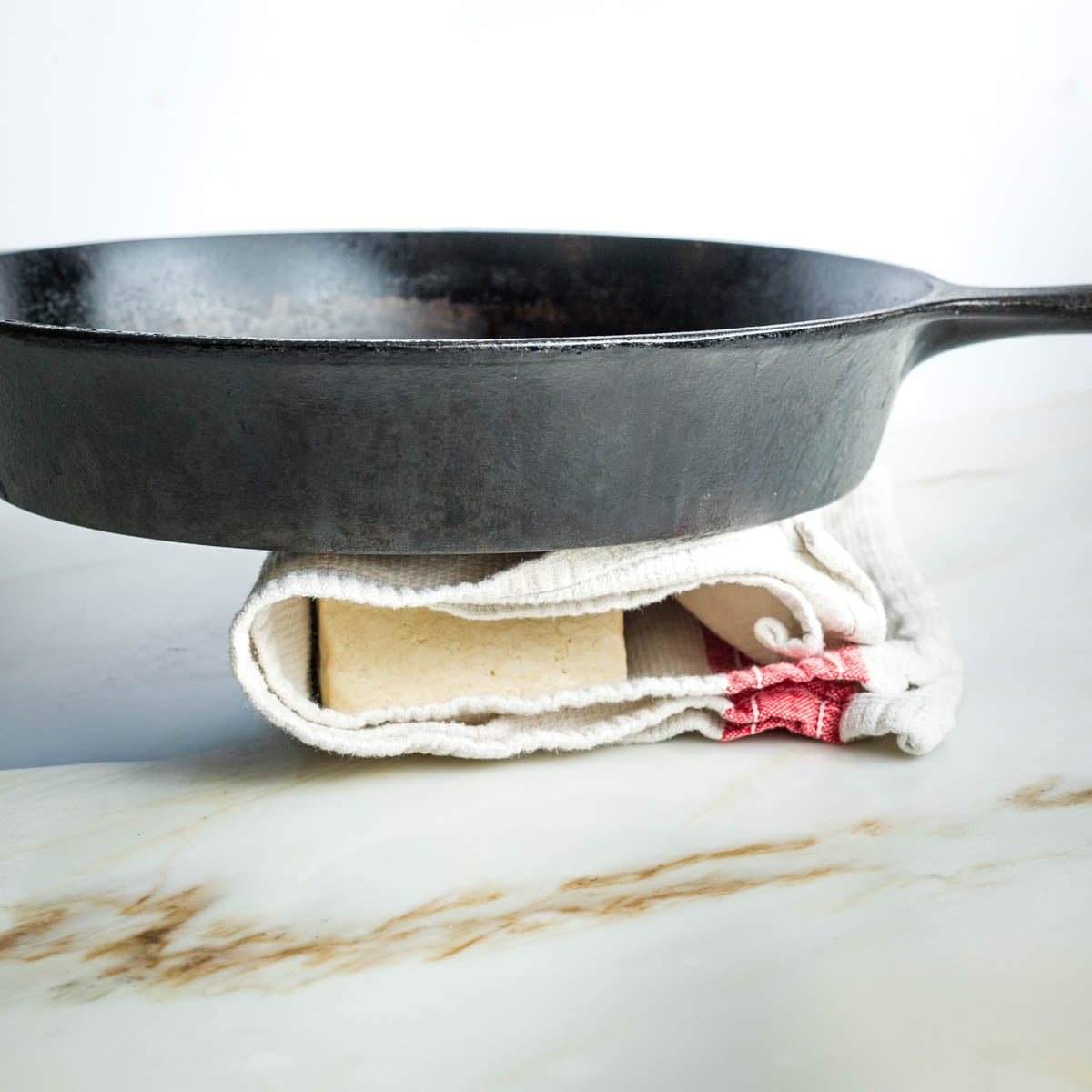 Cast iron skillet on top of a tea towel with block of tofu wrapped inside.