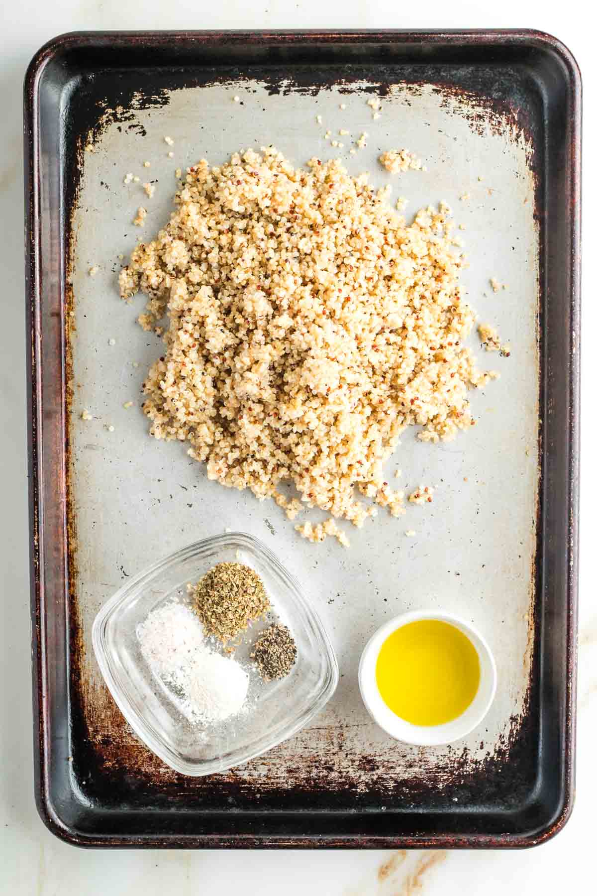 Rimmed baking sheet with a pile of cooked quinoa, a dish of assorted spices and a small dish of olive oil. 