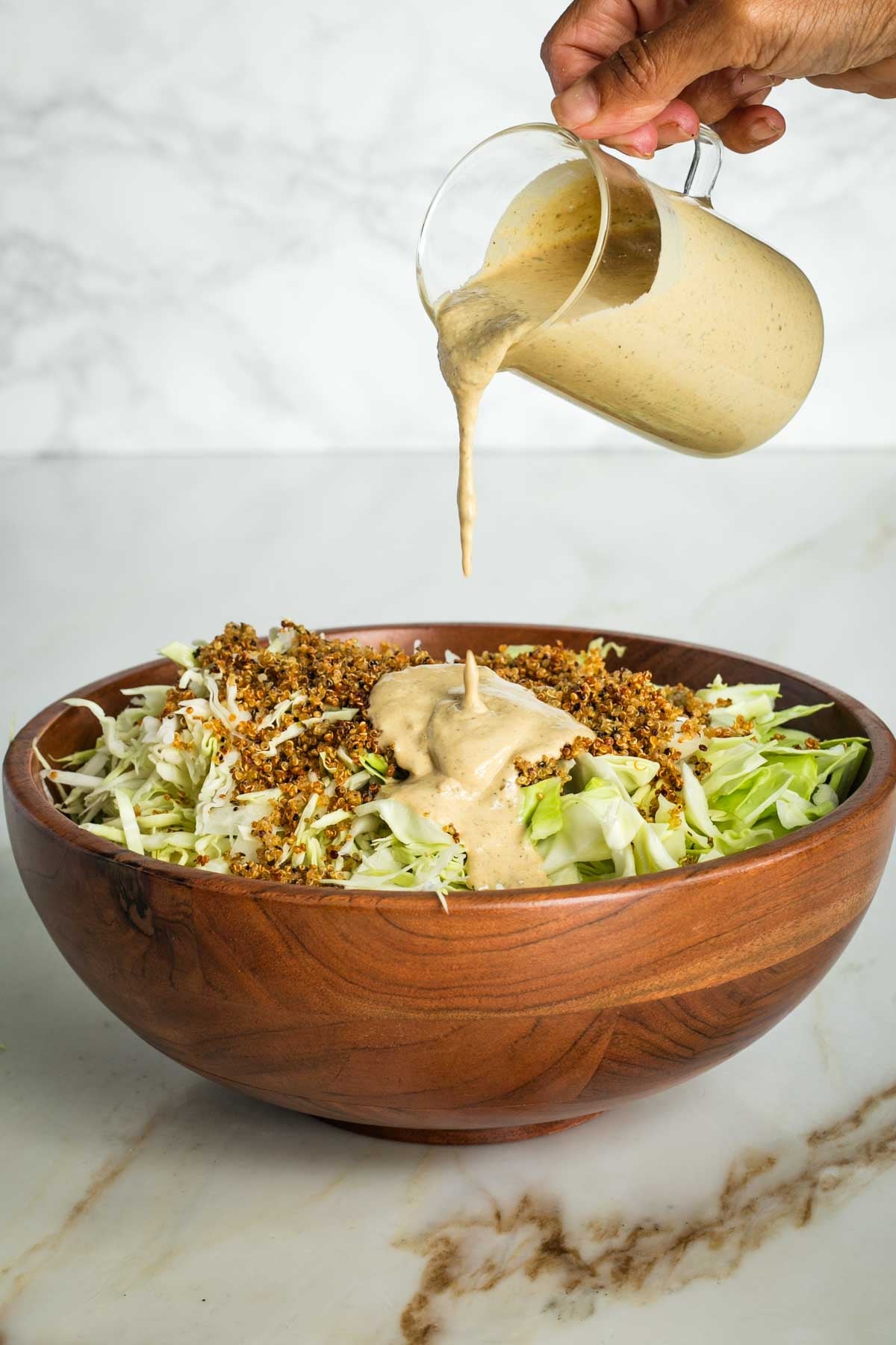 Pouring caesar dressing over a large wooden bowl with chopped cabbage and crispy quinoa.