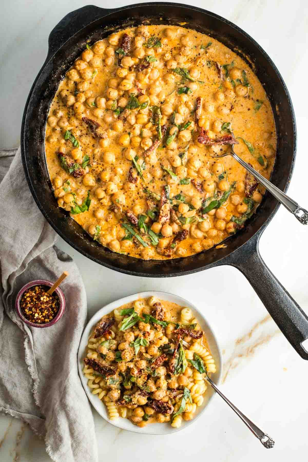 Chickpeas, sun dried tomatoes and fresh basil in a creamy orange sauce in a skillet and then served over pasta on a plate. 