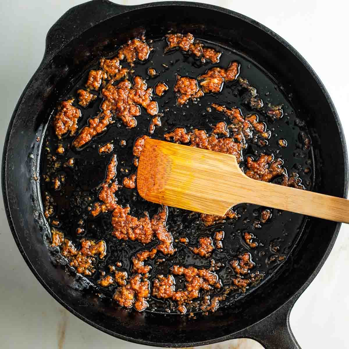Wooden spoon mixing tomato paste into garlic and oil in a cast iron skillet.