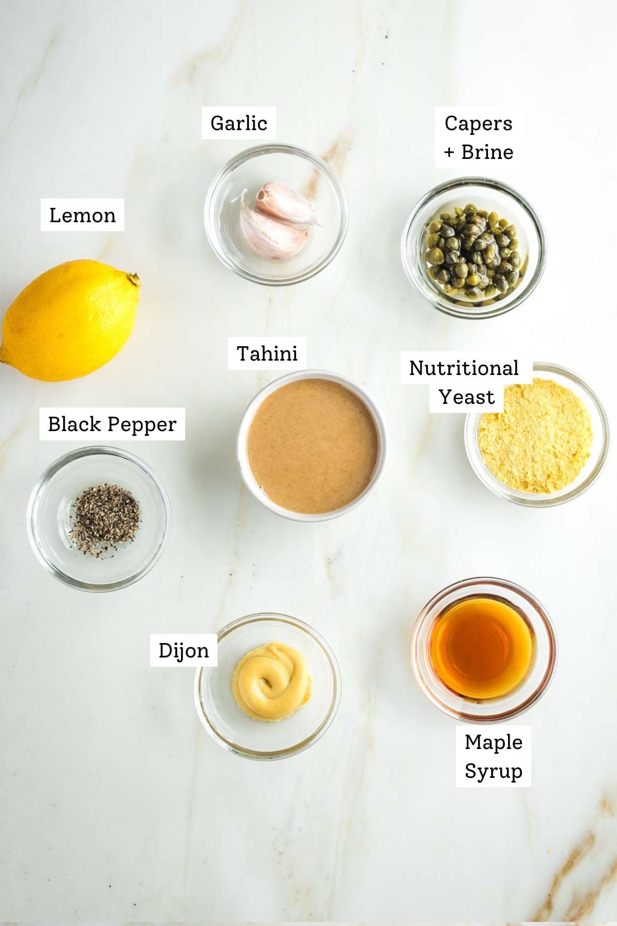 Labeled ingredients for vegan Caesar dressing: tahini, garlic, lemon, capers, nutritional yeast, maple syrup and mustard.
