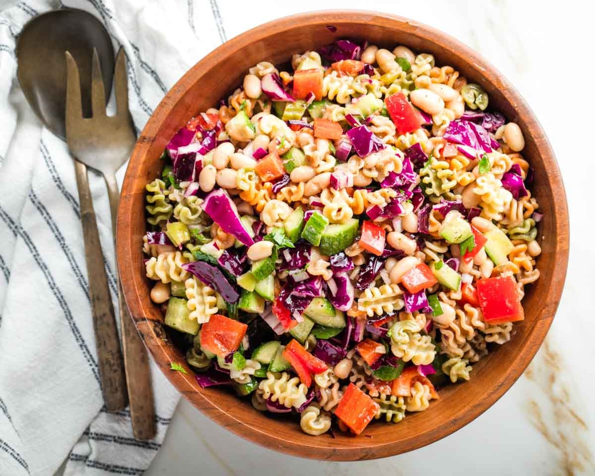 Colorful pasta salad in a wooden bowl with tossers off to the side.