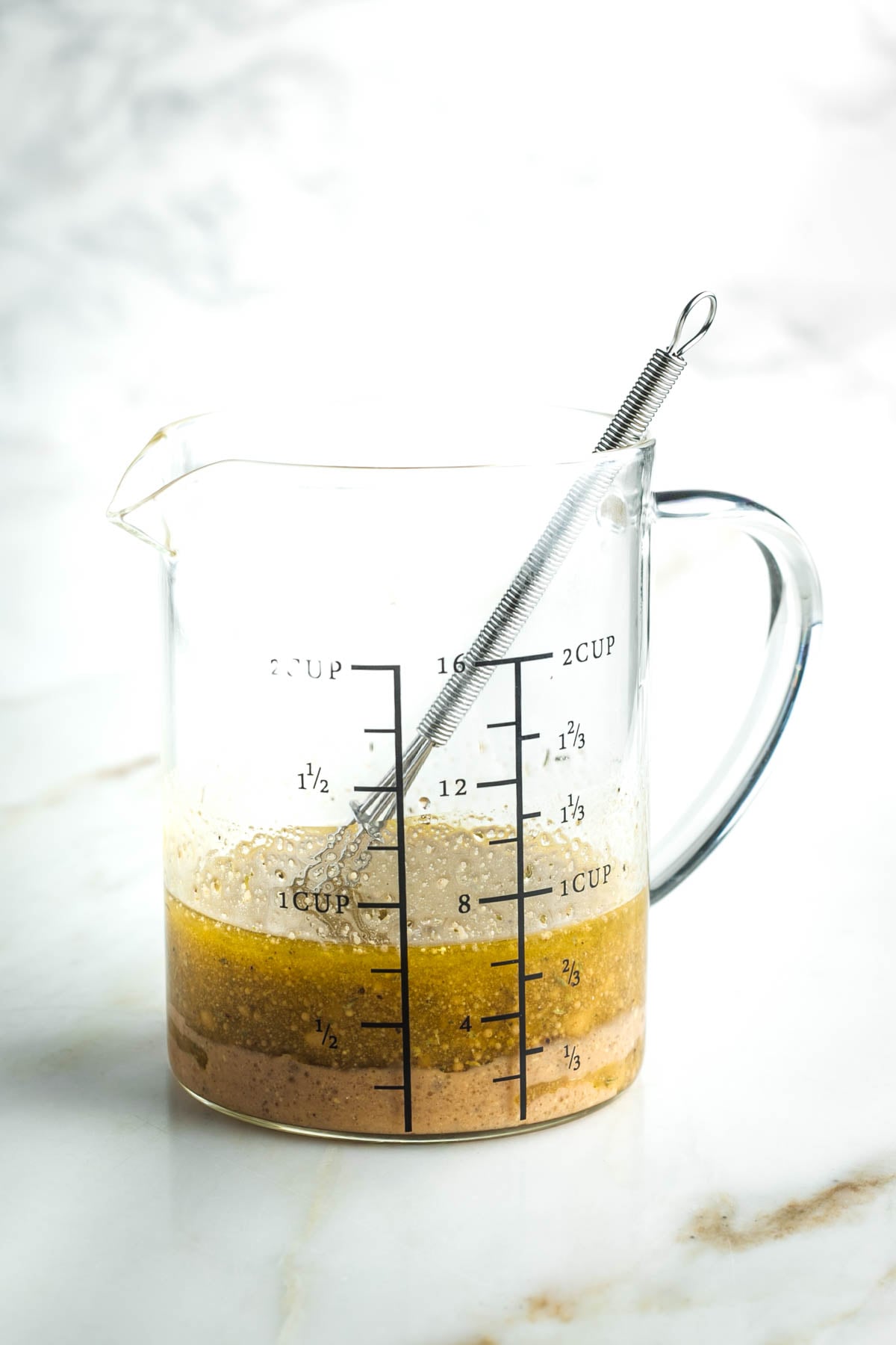 Vinaigrette in a glass pitcher with measurements on the side and a small whisk. 