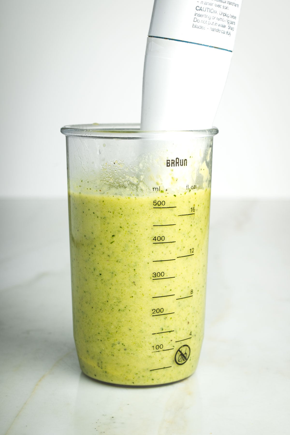 An immersion blender in a tall cup blending a creamy green sauce.