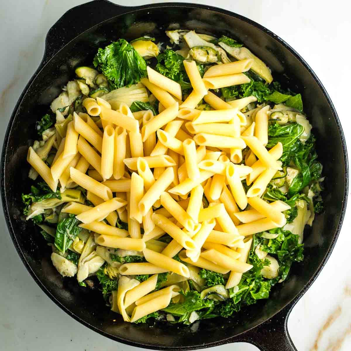 Cooked penne on top of a skillet full of sauteed vegetables.