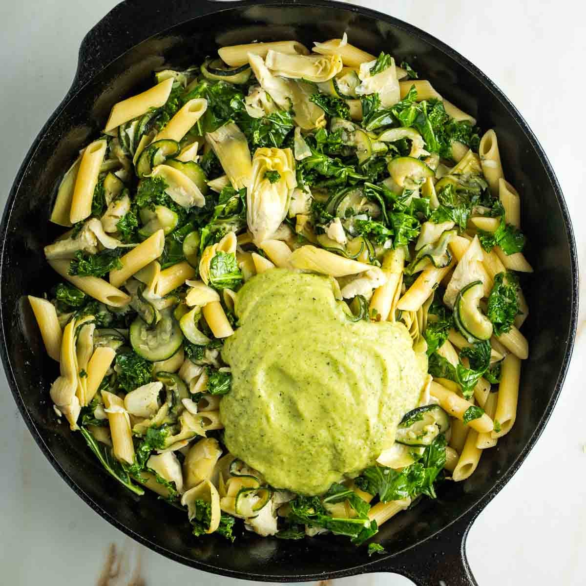 Sauteed zucchini, kale and artichoke hearts with pasta in a skillet and a heap of green creamy sauce.