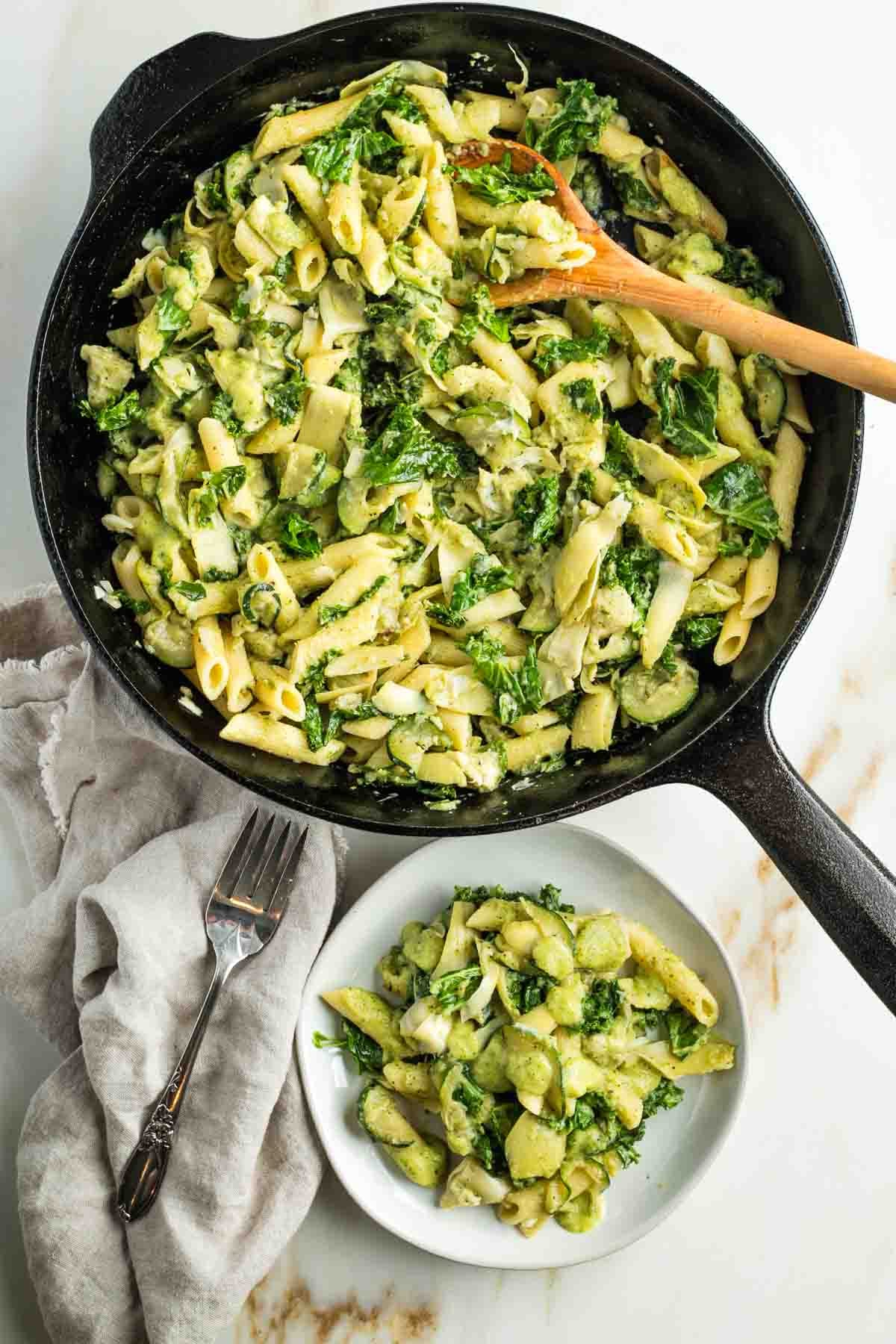 Skillet full of pasta, kale, zucchini and artichoke hearts covered in a thick and creamy green sauce.