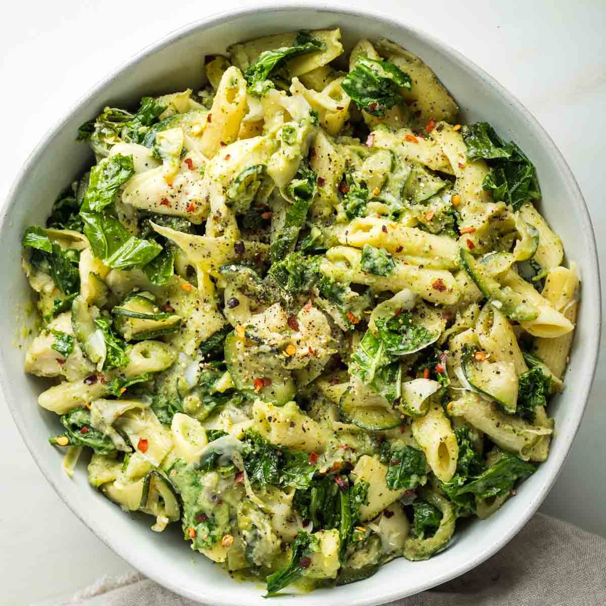 Bowl of pasta with kale, zucchini and artichoke hearts in a creamy green sauce.