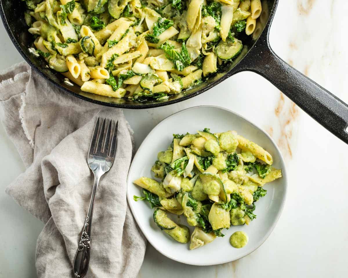 Pasta with zucchini, kale and artichoke hearts in a green creamy sauce on a plate and in a skillet.