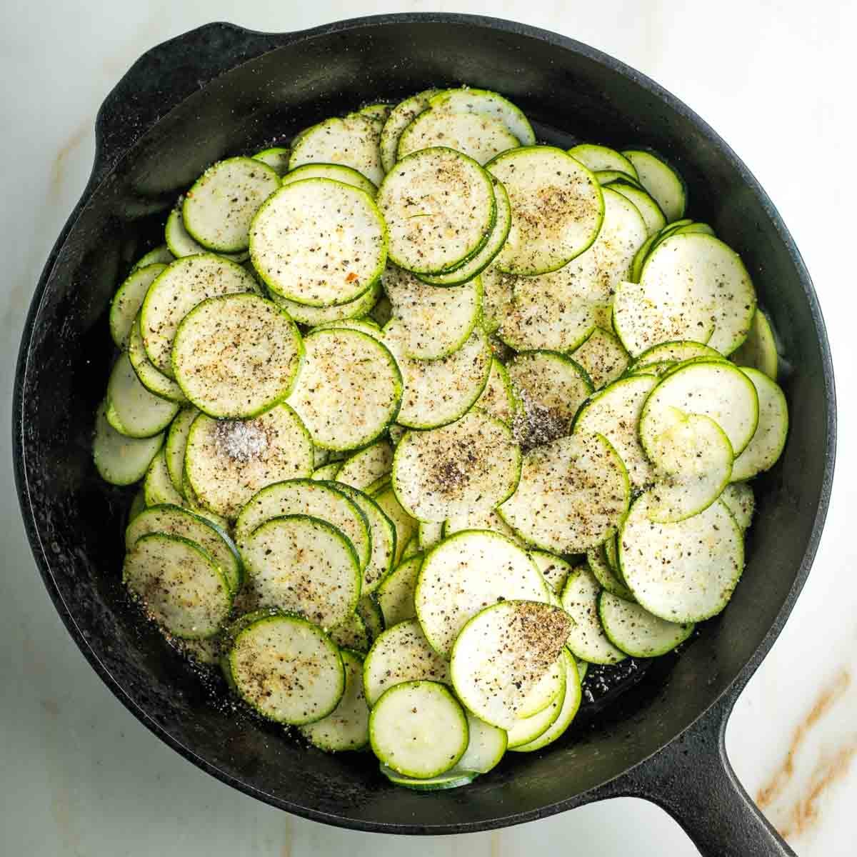 Cast iron skillet filled with thinly sliced zucchini rounds sprinkled with salt and black pepper.