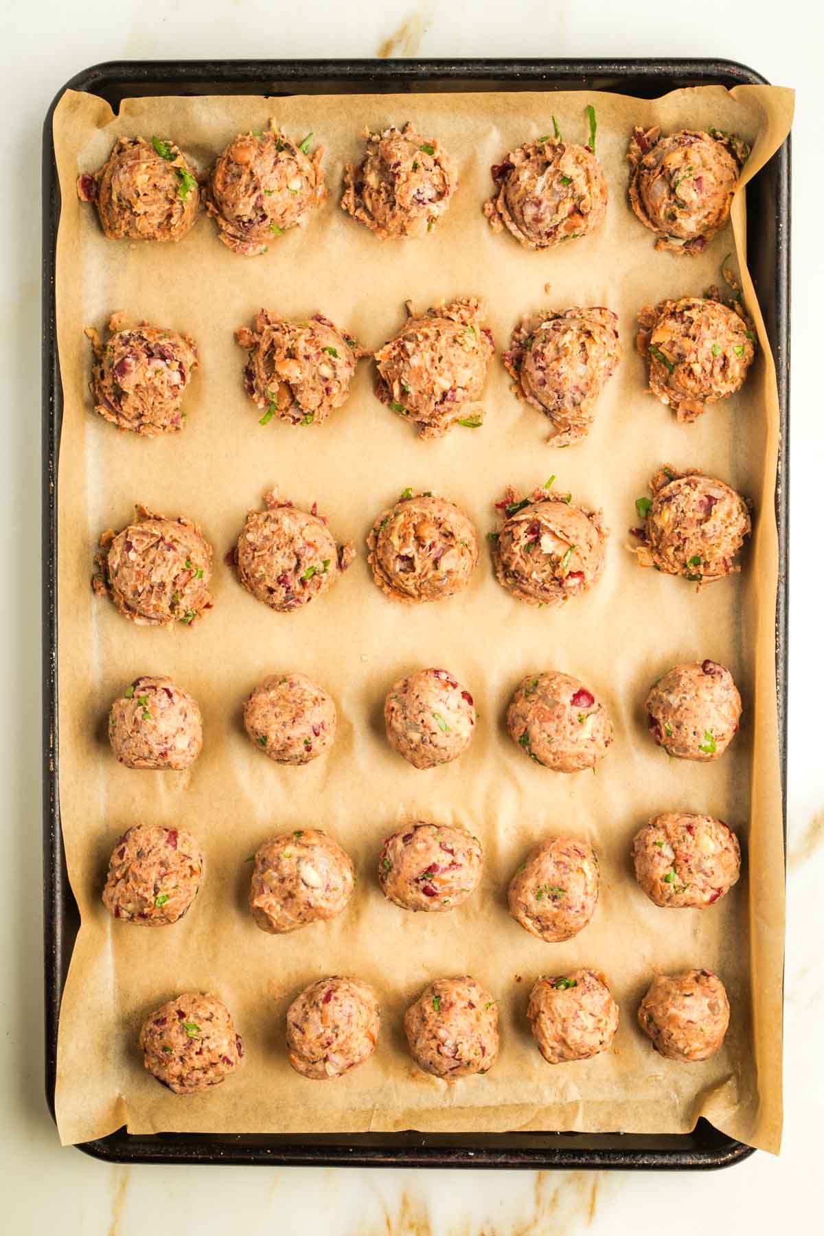 Small vegan Swedish meatballs in heaps on a baking sheet with parchment, half have been rolled into smooth balls.