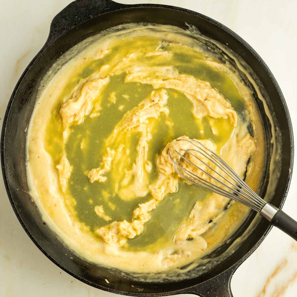 Broth poured onto a roux that's being stirred with a whisk in a cast iron skillet.