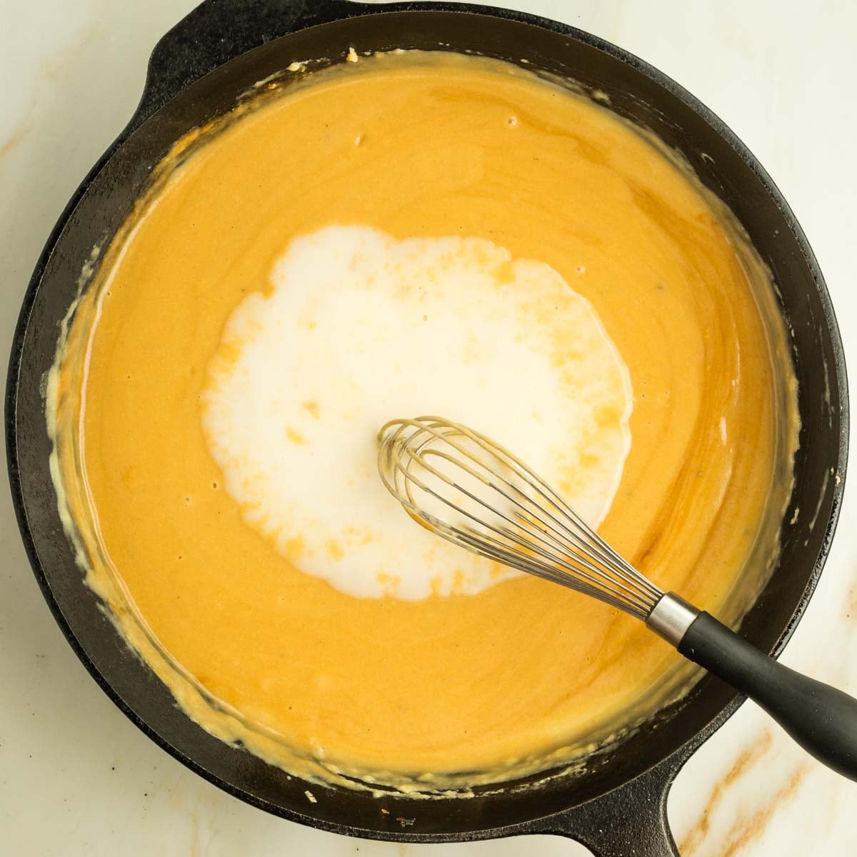 Whisking poured milk into a yellowish brown sauce in a cast iron skillet.