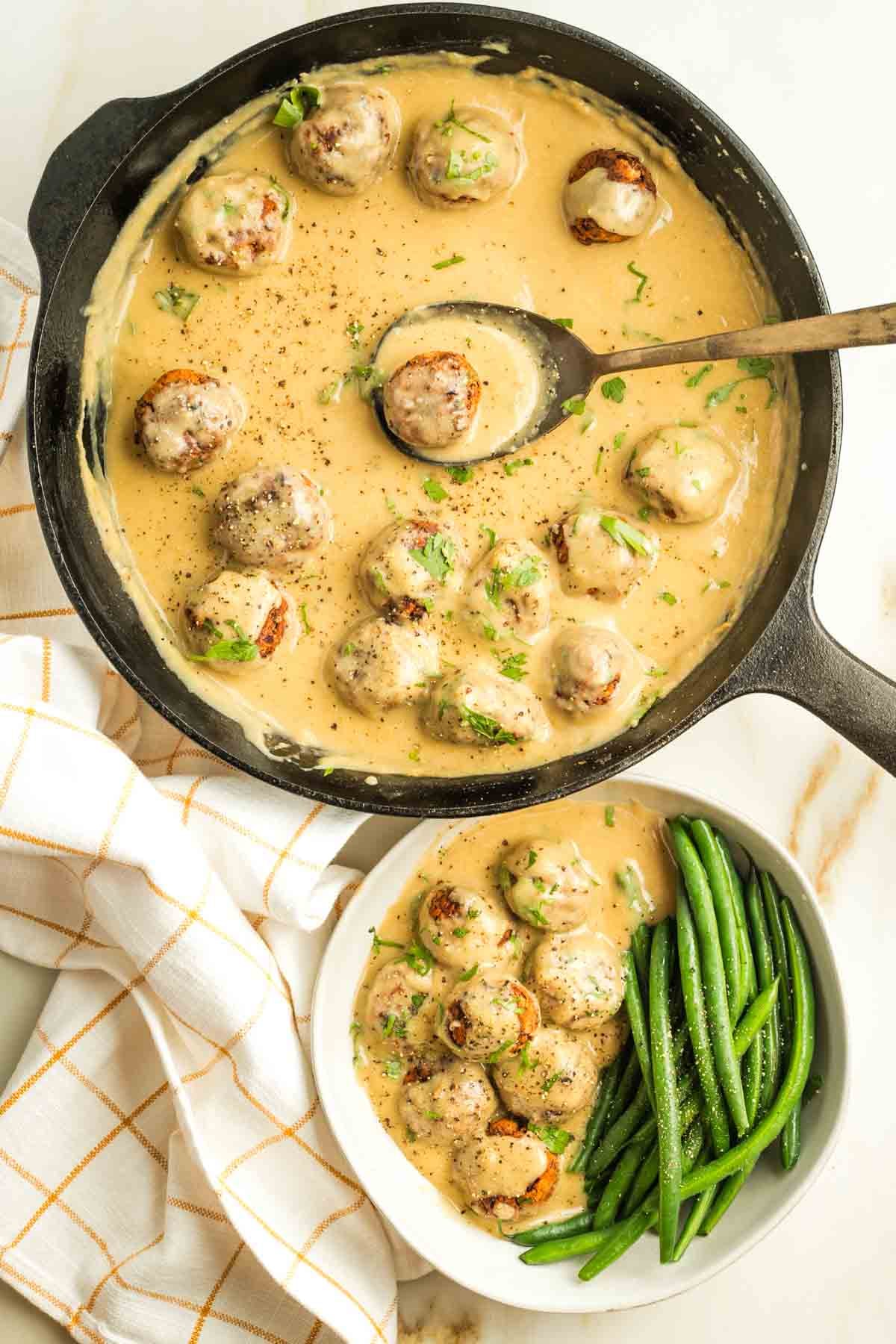 Cast iron skillet with a spoon scooping out a Swedish meatball in sauce and placing into a bowl with green beans and more meatballs in sauce.