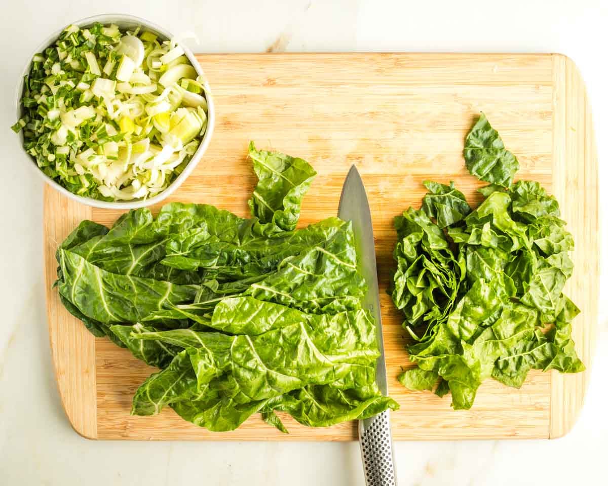 Chopping green chard leaves on a board with a bowl of thinly sliced chard stems and leeks.