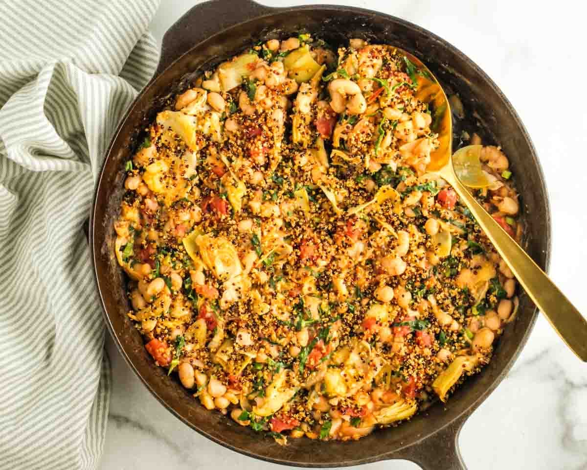 Lemony Tuscan white bean skillet with artichokes and kale, topped with crispy quinoa, in a cast-iron pan with a gold serving spoon.