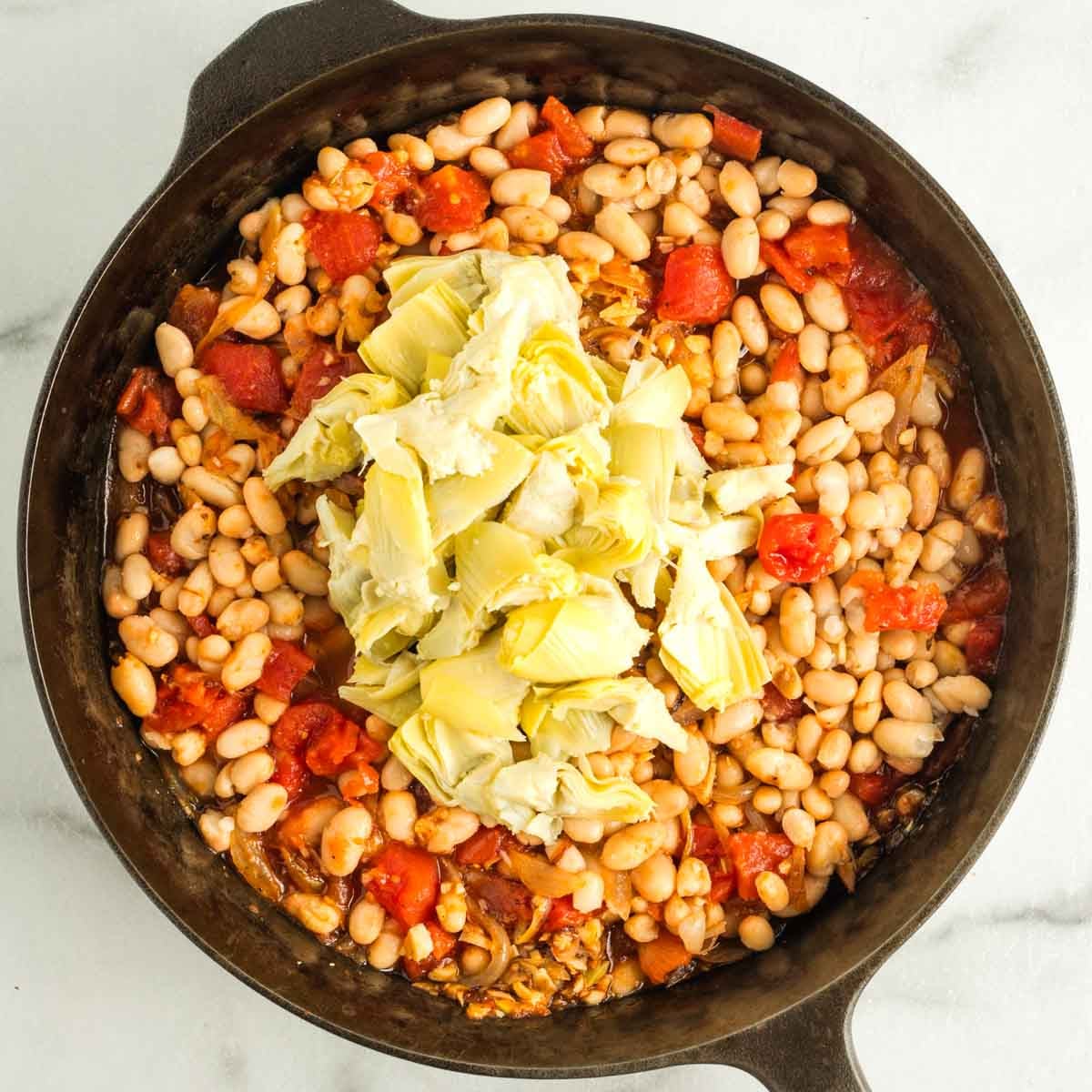 Quartered artichoke hearts on top of beans, tomatoes and onions in a skillet. 