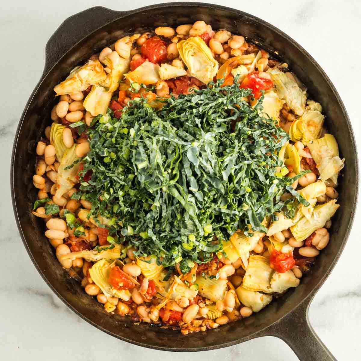 Pile of julienned kale strips on top of a skillet of artichoke hearts, tomatoes, onions and beans.