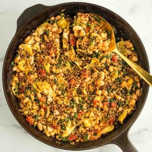 Gold spoon serving up some beans, tomatoes, artichoke hearts from a skillet topped with crispy quinoa.