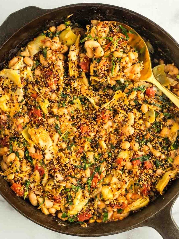 Gold spoon serving up some beans, tomatoes, artichoke hearts from a skillet topped with crispy quinoa.