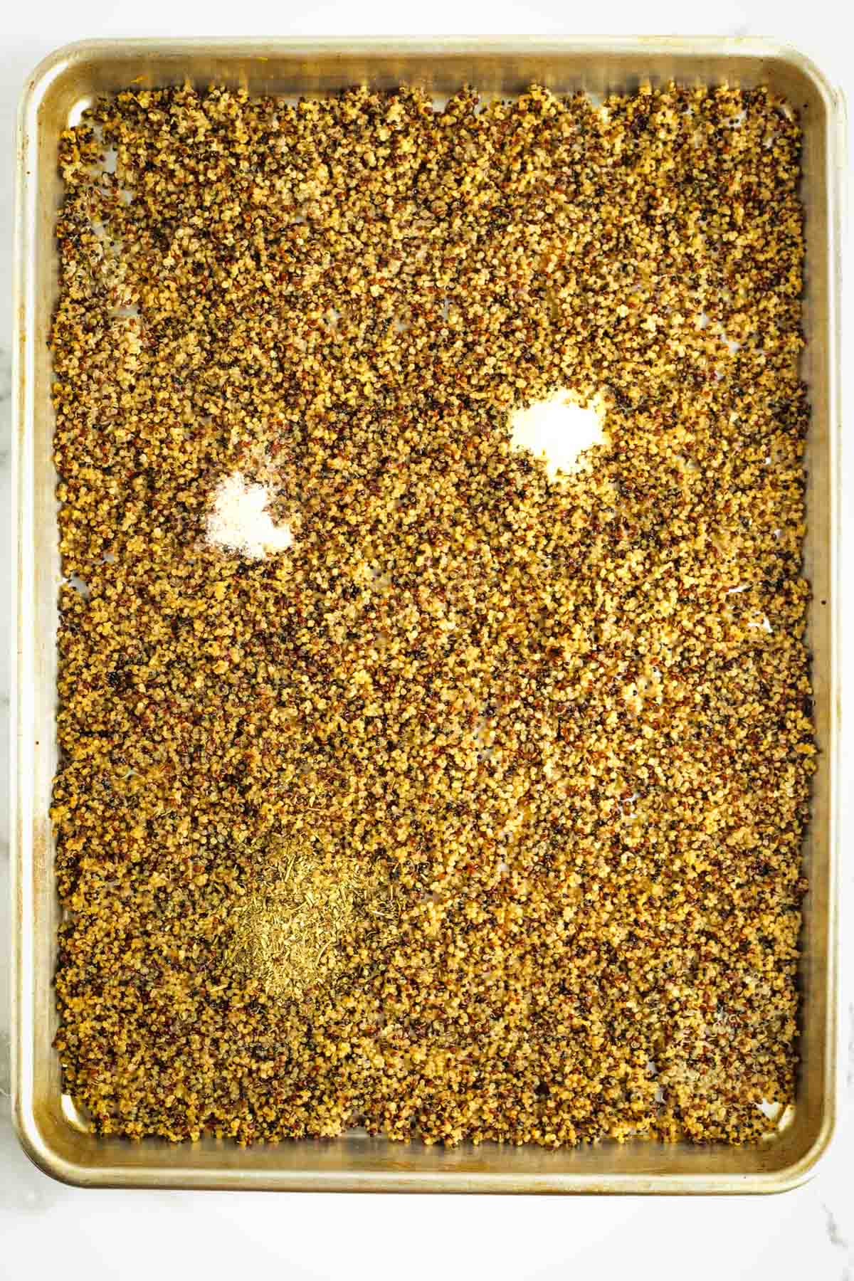 Cooked quinoa spread on a rimmed baking tray with Italian seasoning, salt and garlic powder.